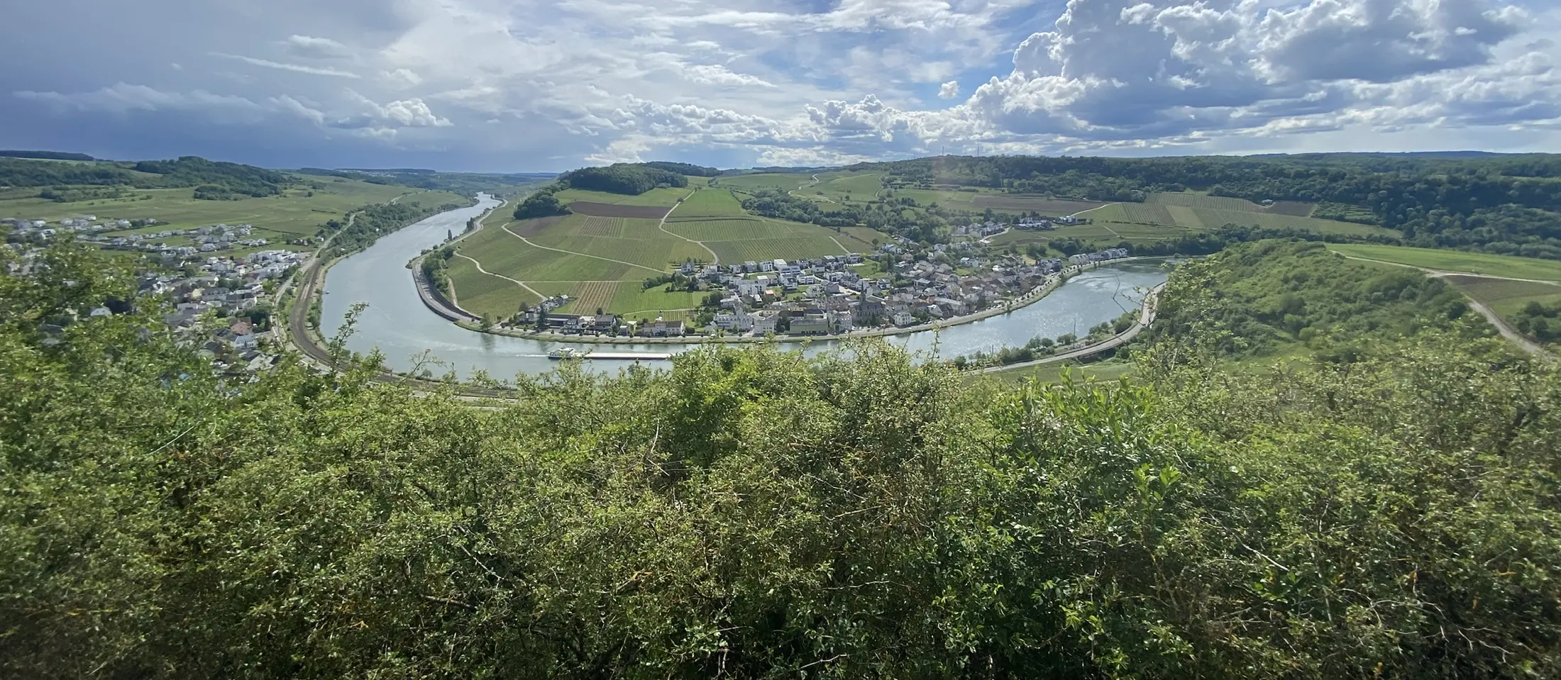 Blick von oben auf eine Moselschleife mit Dorf, Weinbergen und Wolkenhimmel
